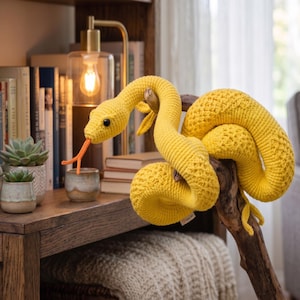 May include: A bright yellow crocheted snake toy with a textured, scale-like pattern, black bead eyes, and a protruding orange forked tongue. The soft, stuffed creature is coiled around a piece of natural driftwood, showcasing its flexible form.