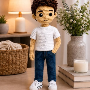 May include: A handmade crochet doll featuring a figure with dark brown hair, a white t-shirt, dark blue jeans, and white sneakers. The doll has a light beige complexion, black button eyes, and a stitched smile, standing upright with one hand in a pocket.