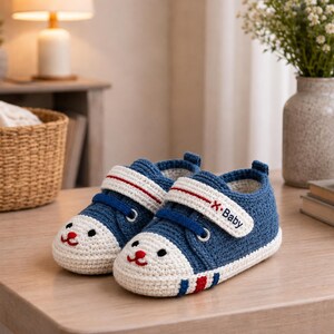 May include: A pair of blue and white crocheted baby sneakers featuring a white toe cap with a small red embroidered face. The shoes have a white hook-and-loop strap with the text X-Baby in black, blue laces, and red and blue stripes on the sole.