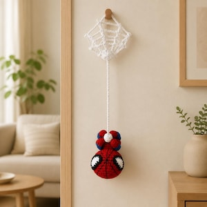 Puede incluir: Un adorno de pared tejido a ganchillo que presenta una telaraña blanca unida a un cordón largo que suspende una cabeza de personaje redonda, roja, azul y negra con grandes ojos blancos. La pieza está hecha de hilo y cuelga de un gancho de madera.