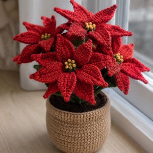 May include: A crocheted poinsettia plant featuring vibrant red petals and small yellow centres, potted in a textured tan crocheted container. The plant has dark green leaves nestled beneath the red blooms, creating a realistic floral display.