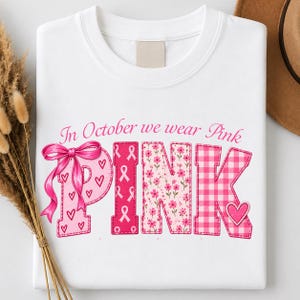 Op de afbeelding: Een wit sweatshirt met ronde hals en de tekst In October we wear Pink. Het woord PINK bestaat uit grote letters met diverse patronen: een P vol hartjes met een roze strik, een I met lintjes, een N met bloemen en een K met een ruitjespatroon.