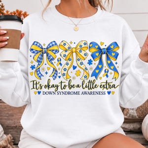 May include: A white crewneck sweatshirt featuring three large decorative bows in blue and yellow patterns. The graphic includes small hearts, flowers, and puzzle pieces, with the text It's okay to be a little extra DOWN SYNDROME AWARENESS.