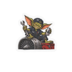 May include: A sticker featuring a green, sharp-toothed creature wearing a black cap with a wrench icon and a dark jumpsuit labeled A&P. The creature holds a metal wrench and a silver can with a jet silhouette, sitting atop a jet engine and red toolbox.