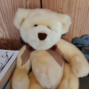 May include: A plush teddy bear with cream-coloured fur, featuring a dark brown stitched nose and a matching brown satin ribbon tied in a bow around its neck. The bear has rounded ears and a soft, fuzzy texture throughout its body and limbs.