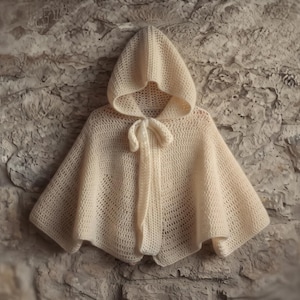 May include: A cream-colored crocheted hooded cape featuring a loose, open-knit texture and a matching tie closure at the neckline. The garment has a flared, poncho-style silhouette that drapes downward from the hood, creating a soft, layered appearance.