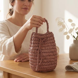 Puede incluir: Un bolso tipo cubo de color rosa empolvado hecho de material tejido, con un cuerpo cilíndrico de superficie texturizada y anudada, una sola asa trenzada y un cierre de cordón a juego que ajusta la parte superior del bolso de forma segura.