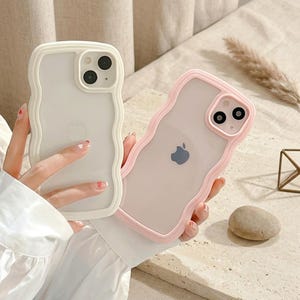Puede incluir: Dos fundas para smartphone con bordes ondulados y festoneados. Una funda es de color crema sólido con una parte trasera translúcida, y la otra es de color rosa claro sólido con una parte trasera translúcida. Ambas tienen un recorte cuadrado.