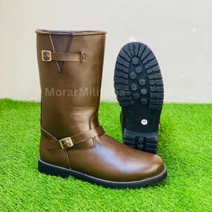 May include: A brown leather mid-calf boot featuring two adjustable straps with gold-toned buckles, a dark brown elastic side panel, and a thick black rubber lug sole. A second boot is positioned behind it, showing the black treaded sole.