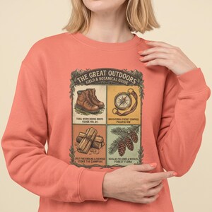 May include: A coral-colored crewneck sweatshirt featuring a graphic print titled The Great Outdoors Field & Botanical Guide. The design includes four panels depicting brown hiking boots, a pocket compass, stacked firewood, and pine cones with needles.