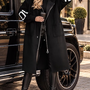 May include: A black hooded long coat featuring a silver zipper closure and two front pockets. The sleeves display white text reading 10 on the right and 13 on the left. The coat is worn over a black top and black pants with black boots.