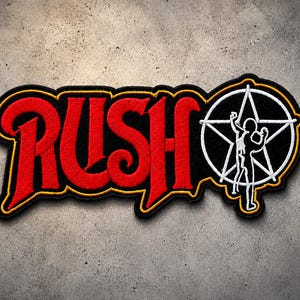 May include: An embroidered patch featuring the word RUSH in bold red lettering with a yellow border. To the right, a white star-man logo is enclosed within a white circle on a black background, all outlined in yellow thread on a black fabric base.