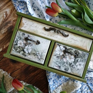 Op de afbeelding: Een groene houten bureau-organizer met drie lades. De bovenste lade is voorzien van cursieve tekst, terwijl de twee onderste lades een wit bloemenpatroon hebben. Elke lade is uitgerust met een decoratieve handgreep van donker metaal.