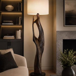 May include: A floor lamp featuring a tall, dark brown, organic-shaped wooden base with a central cutout, standing on a circular black base. The lamp is topped with a large, cylindrical, off-white fabric lampshade that emits a warm, soft glow.