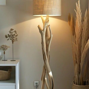 May include: A floor lamp featuring a base constructed from multiple intertwined pieces of light-coloured natural driftwood. The lamp is topped with a cylindrical drum lampshade in a neutral beige fabric, emitting a warm glow from the light source.