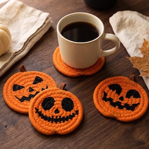 May include: Three round, orange, woven rope coasters shaped like pumpkins, each featuring a unique black stitched jack-o'-lantern face. The coasters are crafted from coiled orange cord with a small brown fabric tab at the top of each pumpkin.