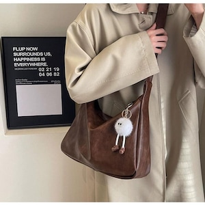 May include: A dark brown hobo-style shoulder bag with a smooth finish and a wide strap. A white fuzzy pom-pom charm with dangling legs and small brown shoes is attached to the bag's zipper. The bag is worn over a beige coat.