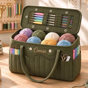 May include: An olive green quilted yarn storage bag with two handles, featuring the embroidered name Cmnia and two small flowers. The open lid displays various crochet hooks and needles, while the main compartment holds eight colourful yarn balls.