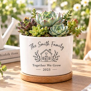 Puede incluir: Una maceta cilíndrica de cerámica blanca con base de madera, llena de varias suculentas verdes y moradas. La maceta presenta texto en negro que dice The Smith Family, Together We Grow, 2025 y un gráfico de una casa con un corazón encima.