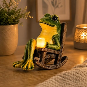 Può includere: Una statuetta di rana verde e gialla seduta su una sedia a dondolo in legno marrone. La rana tiene in mano una piccola candela cilindrica bianca luminosa. La statuetta ha grandi occhi neri e una consistenza liscia con una luce calda emanata.