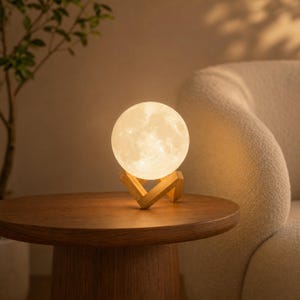Peut inclure: Une lampe lune sphérique émettant une lueur chaude et douce, dotée d'une surface texturée imitant le paysage lunaire. La lampe est soutenue par un support en bois clair composé de deux pièces imbriquées formant une base stable.