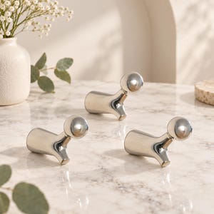 Op de afbeelding: Drie gepolijste zilverkleurige metalen wandhaken, elk met een gladde, cilindrische basis die naar buiten uitloopt en eindigt in een afgeronde, bolvormige knop aan de bovenkant. De haken hebben een reflecterende afwerking en een gebogen vorm.