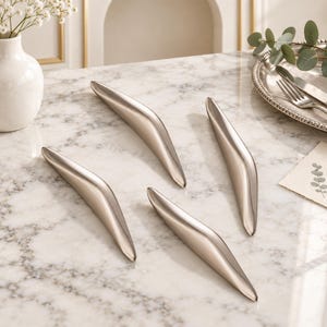 May include: Four sleek, curved cabinet pulls with a polished silver-tone metallic finish. Each handle features a smooth, elongated, and tapered design with a reflective surface, arranged in a scattered pattern on a white marble surface.