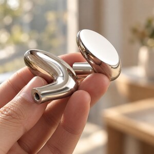 May include: Two polished silver-toned metal cabinet knobs with a smooth, reflective finish. One knob features a rounded, circular face, while the other has an elongated, curved, and tapered shape with a visible threaded screw hole at the base.