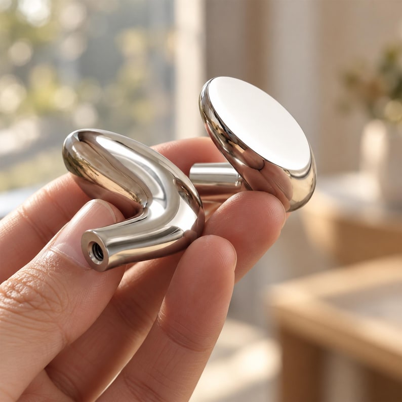 May include: Two polished silver-toned metal cabinet knobs with a smooth, reflective finish. One knob features a rounded, circular face, while the other has an elongated, curved, and tapered shape with a visible threaded screw hole at the base.