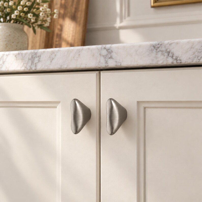 May include: Two brushed silver cabinet knobs with a smooth, rounded triangular shape. These metal hardware pieces are mounted on off-white cabinet doors featuring a recessed panel design, providing a clean and functional look for cabinetry.