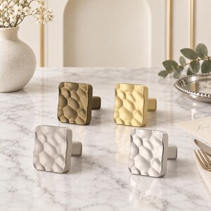 May include: Four square cabinet knobs with a hammered, dimpled texture. The set includes two knobs with a brushed brass finish, one knob with a matte silver finish, and one knob with a polished chrome finish, each featuring a central mounting post.