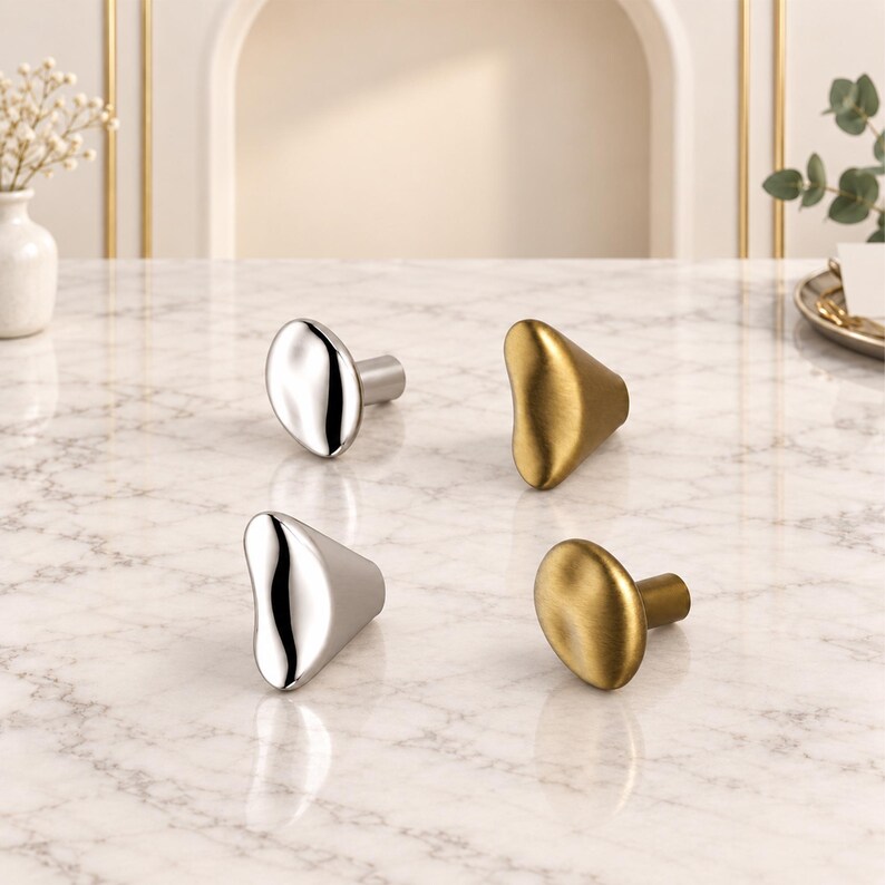 May include: Four cabinet knobs arranged on a surface. Two knobs have a polished silver finish with irregular, organic shapes, while the other two feature a matte gold finish with smooth, rounded, and slightly asymmetrical pebble-like forms.
