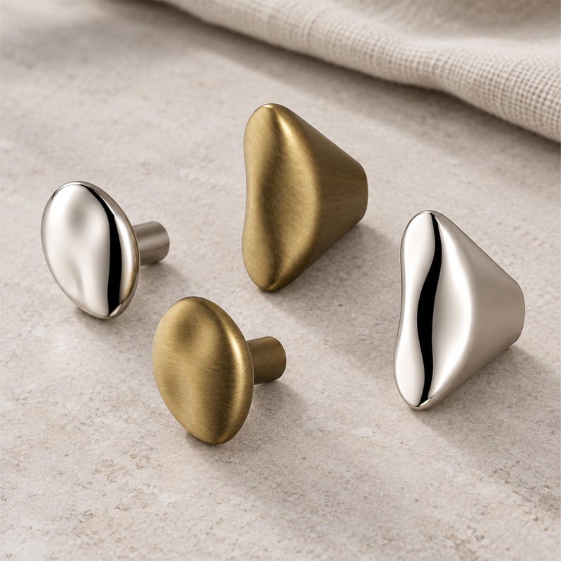 May include: A collection of four cabinet knobs featuring irregular, organic shapes. Two knobs have a polished, reflective silver finish, while the other two knobs have a brushed, matte gold finish. Each knob includes a short cylindrical mounting post.