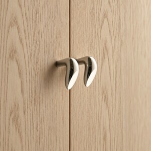 May include: Two polished chrome cabinet knobs with a curved, teardrop-like shape mounted on light-colored wood cabinet doors. The metal hardware features a smooth, reflective surface and a distinct angled design that protrudes from the wood grain.