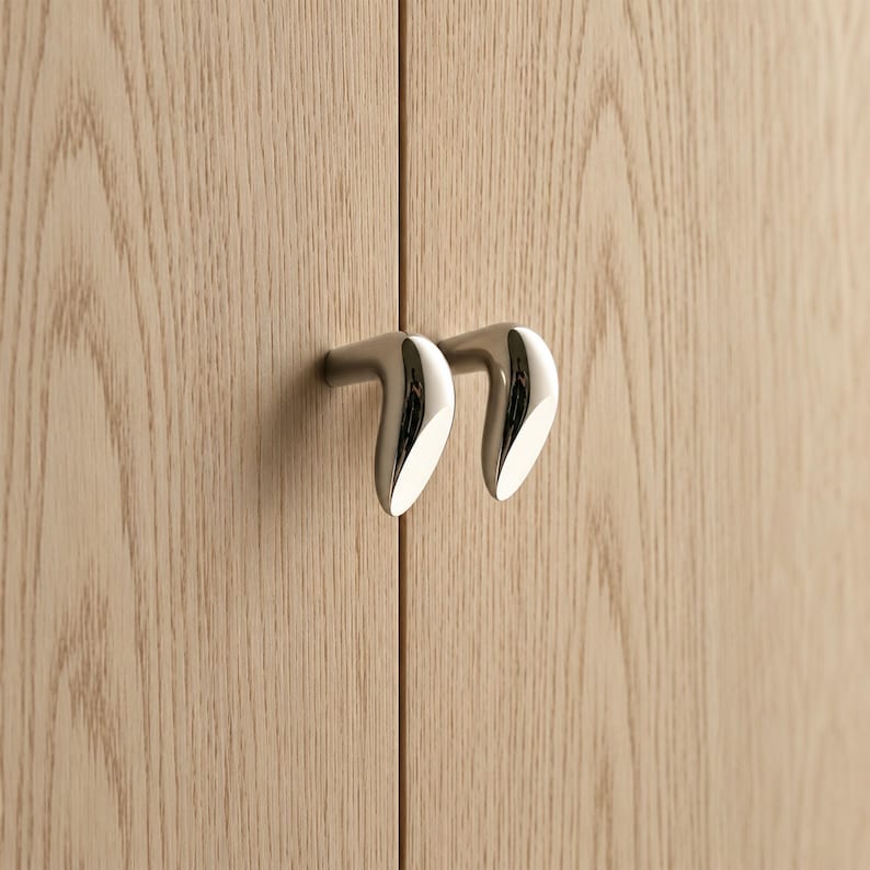May include: Two polished chrome cabinet knobs with a curved, teardrop-like shape mounted on light-colored wood cabinet doors. The metal hardware features a smooth, reflective surface and a distinct angled design that protrudes from the wood grain.