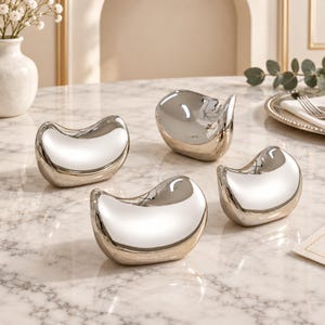 May include: Four crescent-shaped, silver-toned metallic objects with a smooth, reflective, mirror-like finish. Each piece features a curved, concave top surface and a rounded base, designed as individual decorative accents or functional holders.