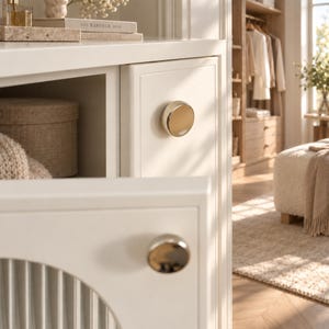 Pode incluir: Um arm&aacute;rio branco com dois puxadores circulares de metal dourado polido. A porta do arm&aacute;rio tem um design de painel vertical canelado. O arm&aacute;rio est&aacute; posicionado numa sala com ch&atilde;o de madeira, um tapete bege e um cabideiro ao fundo.