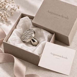 May include: A silver-toned metal knob with a textured, irregular circular top and a smooth, tapered base, nestled inside a beige cardboard gift box lined with branded tissue paper. The box lid and a small square card both feature the text Angusmacleods.