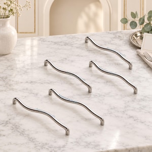 May include: A set of five silver-toned metal cabinet pulls, each featuring a distinct wavy, serpentine silhouette with a smooth, polished finish. These hardware handles are designed for attachment to furniture doors or drawers, showcasing a curved, elongated form that provides a comfortable grip for opening and closing cabinetry.
