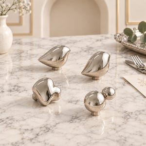 May include: A set of four decorative cabinet knobs with a polished silver finish. Two knobs are shaped like stylised birds, one is an abstract organic form, and one consists of two connected spheres. Each knob features a small cylindrical base.