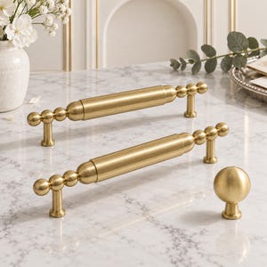 May include: Two brushed brass cabinet pulls featuring a central cylindrical bar flanked by spherical accents and two vertical supports, accompanied by a single matching spherical cabinet knob with a small circular base.