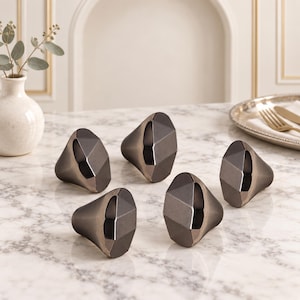 Op de afbeelding: Een set van vijf donkere metalen kastknoppen met een gefacetteerde, geometrische, diamantachtige vorm. Elke knop heeft een glad, meervoudig hoekig oppervlak met een gepolijste afwerking die het licht reflecteert.