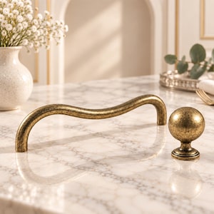 May include: A curved, wavy cabinet handle and a matching spherical cabinet knob, both featuring a distressed, antique brass finish with dark, textured speckles throughout their metallic surfaces, displayed together on a white marble surface.