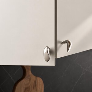 May include: Two polished silver-toned metal cabinet knobs attached to light cream-colored cabinet doors. One knob is oval-shaped and mounted on the front, while the other is a curved, L-shaped handle mounted on the side of the adjacent cabinet door.