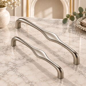 Könnte beinhalten: Zwei Schrankgriffe aus gebürstetem Nickel mit einem glatten, länglichen Bogendesign. Jeder Griff verfügt über einen mittig leicht verbreiterten Bereich, der zu den abgerundeten Enden hin schmaler wird und ein elegantes Metallfinish bietet.
