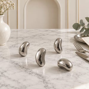 May include: Four small, polished silver-toned metal objects with a smooth, curved, bean-like shape. These reflective, solid pieces are arranged on a surface, showcasing a high-gloss finish that captures light and creates distinct highlights.