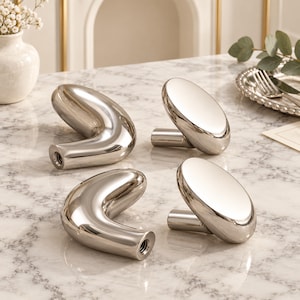 May include: A set of four silver-toned metal cabinet knobs featuring two distinct shapes: two curved, hook-like designs and two smooth, oval-faced knobs. Each piece has a polished, reflective finish and a threaded mounting point at the base.