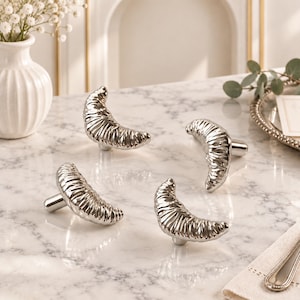 May include: A set of four silver-toned cabinet knobs, each shaped like a crescent moon with a textured, ribbed surface. These metallic hardware pieces feature a polished finish and a cylindrical base for mounting onto furniture or cabinetry doors.