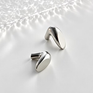 May include: Two silver-toned metal cabinet knobs with a smooth, polished finish. Each knob features a curved, L-shaped design with a rounded, elongated head that tapers slightly, providing a sleek and reflective surface for furniture hardware.