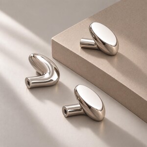 May include: Three polished silver-toned metal cabinet knobs with a smooth, rounded, and organic shape. Each knob features a cylindrical base with a threaded hole for mounting, showcasing a reflective, high-gloss finish and a curved, pill-like head.